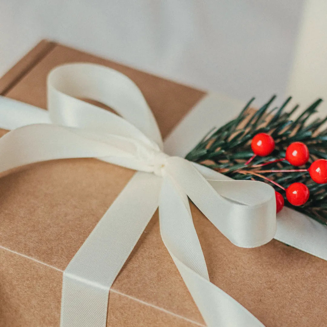 A gift box wrapped in kraft paper with a white satin bow, with a pine sprig and red berries
